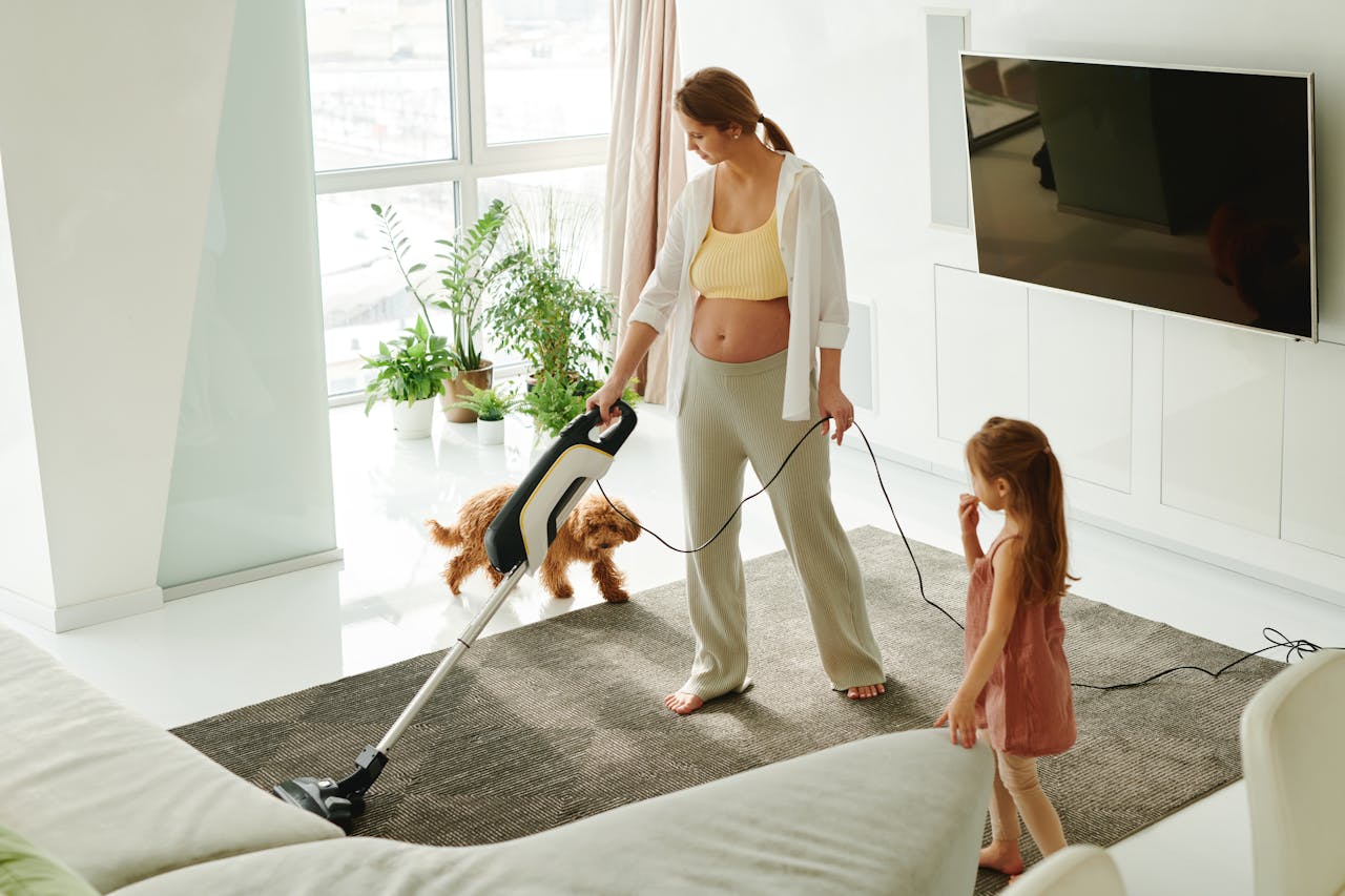 who-we-are Pregnant mother cleaning carpet with vacuum as daughter and dog play indoors.